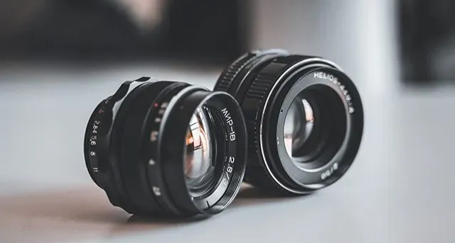 Two camera lenses on a surface, one black and one silver, against a blurred background