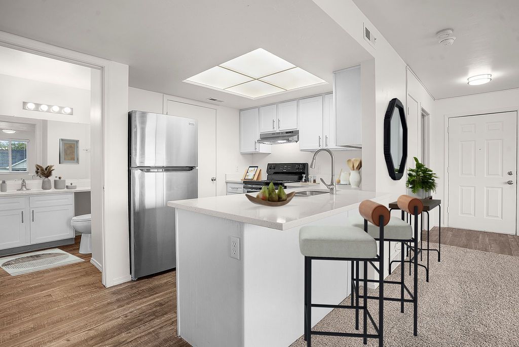 Bright white kitchen with stainless fridge, island seating, and open doorway to a bathroom at Creekside Village apartments in Sandy, UT.