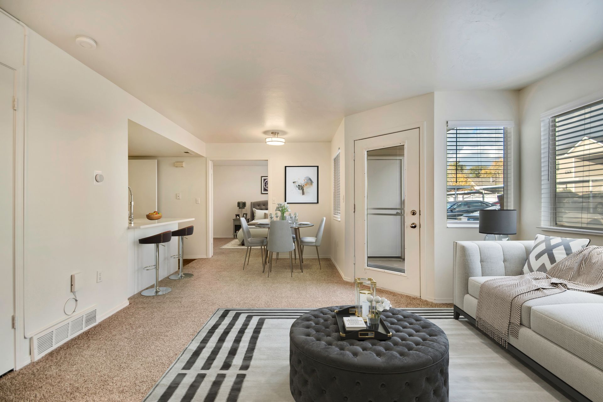 Bright modern living room with beige sofa, striped rug, ottoman, windows, and dining area in the background at Creekside Village apartments in Sandy, UT.