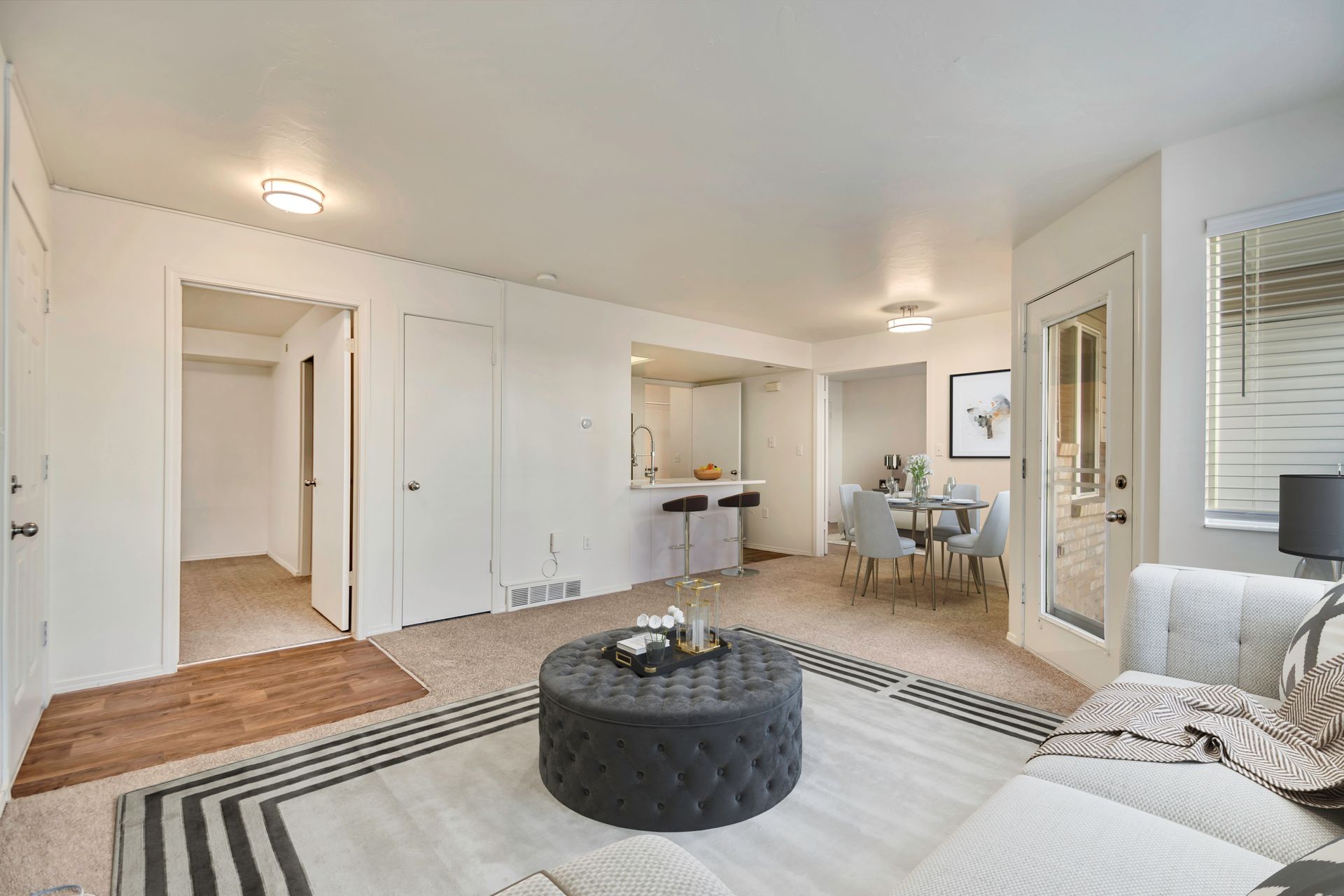 Bright living room with white walls, striped rug, black ottoman, and dining area in back at Creekside Village apartments in Sandy, UT.