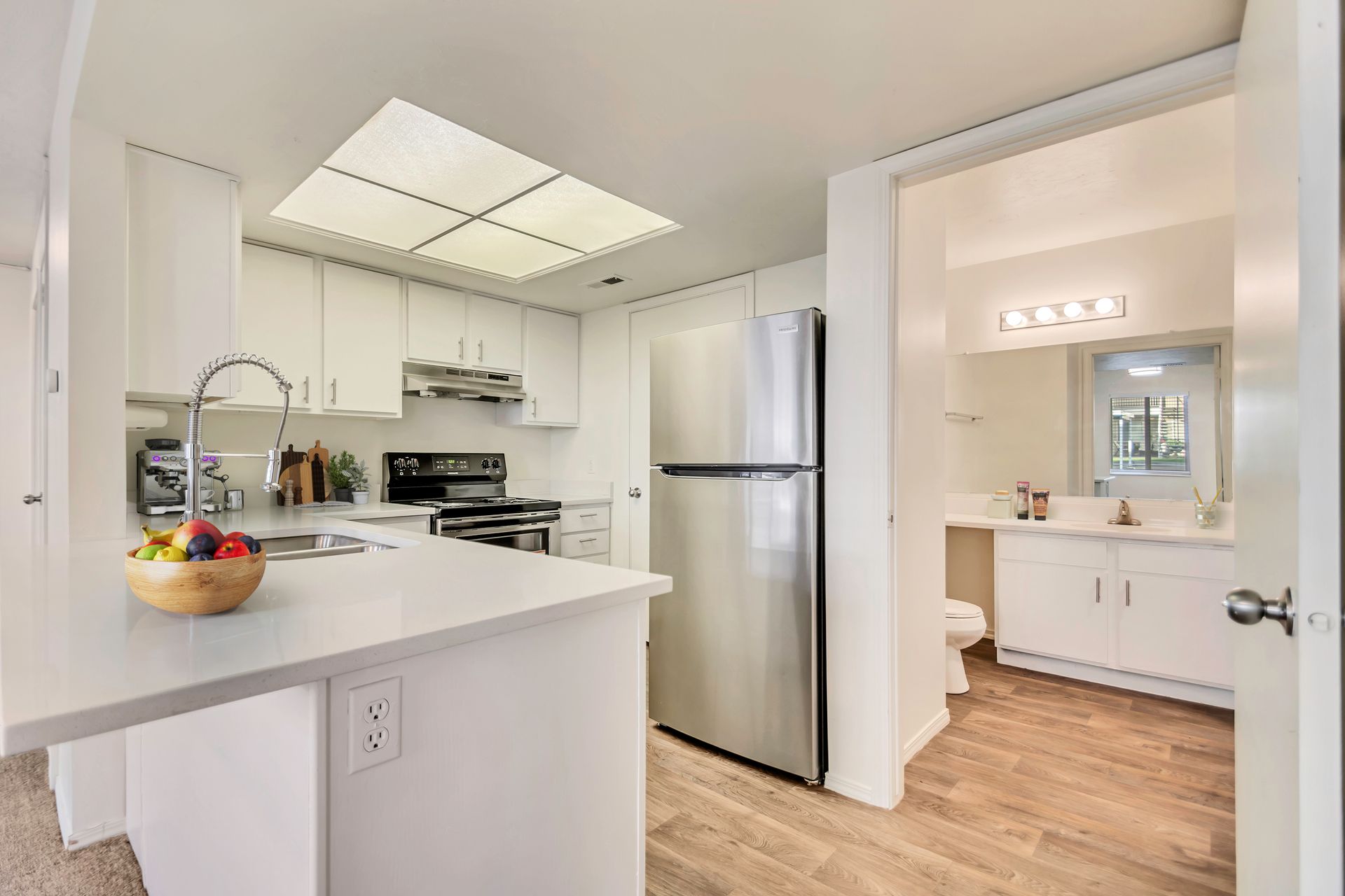 Bright kitchen with stainless steel fridge, white counters, and an adjacent bathroom doorway at Creekside Village apartments in Sandy, UT.
