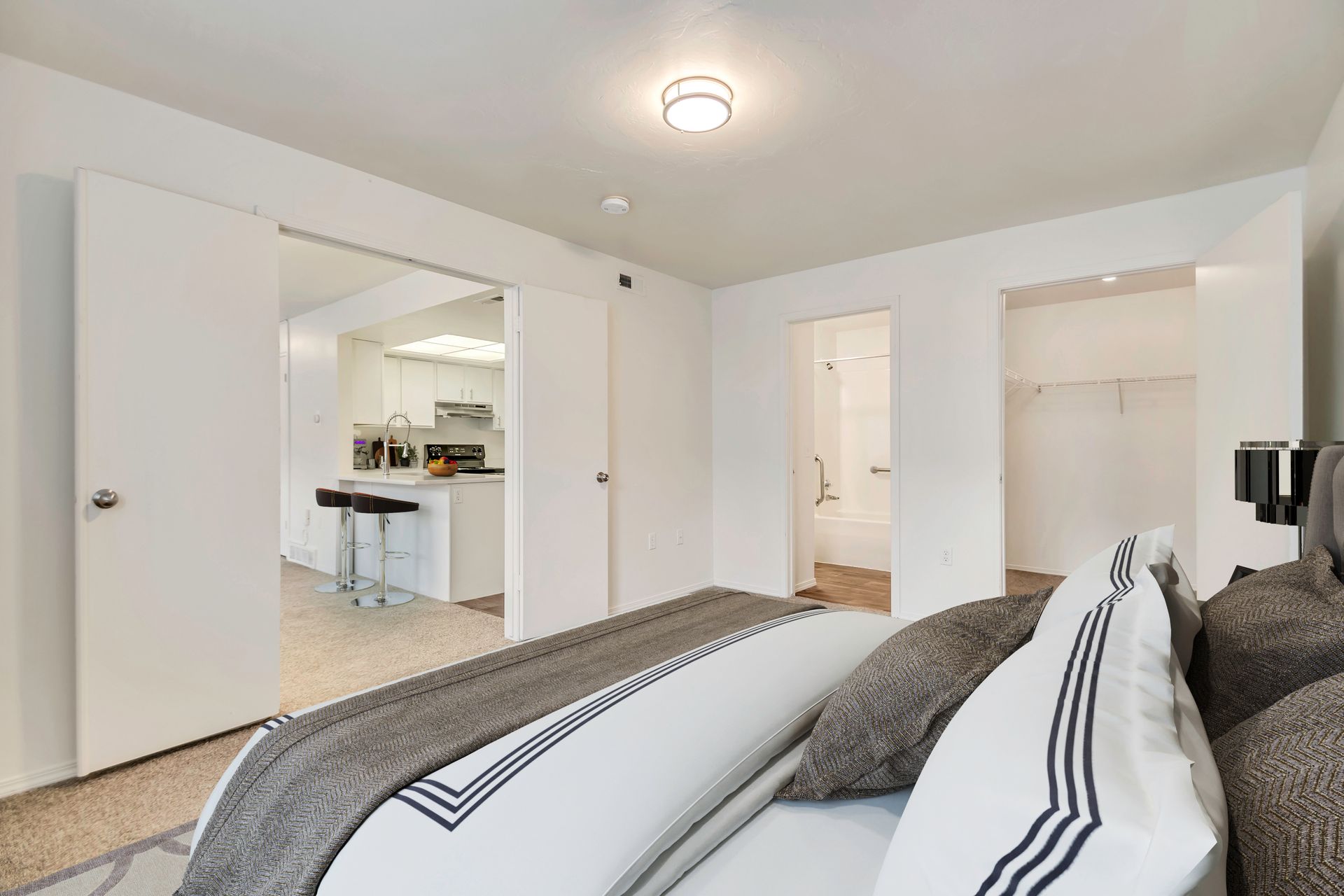 Bright bedroom with white walls, open doors, and a view into a kitchen area at Creekside Village apartments in Sandy, UT.
