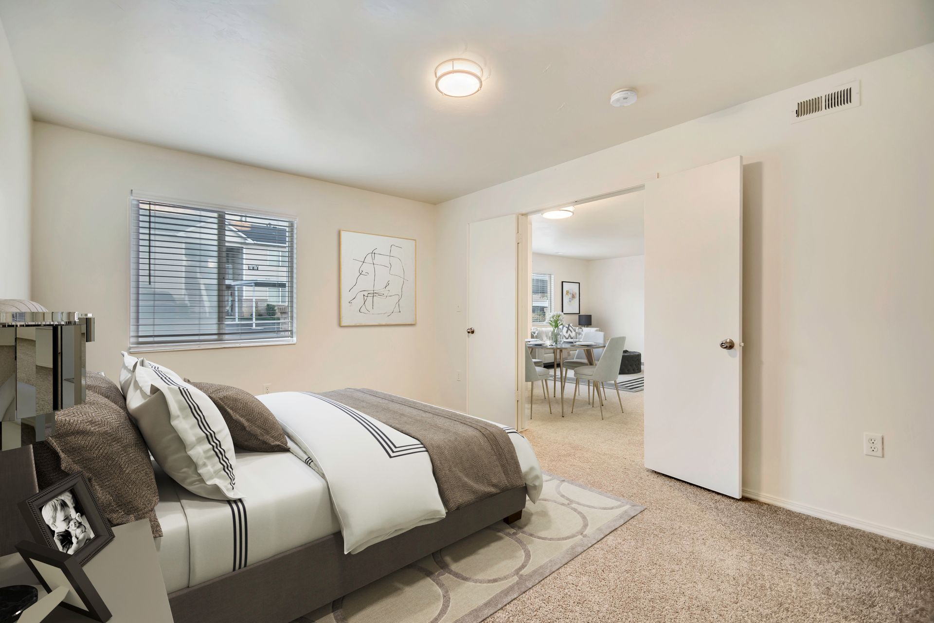 Bright bedroom with a bed, window, wall art, and open door to a dining area at Creekside Village apartments in Sandy, UT.