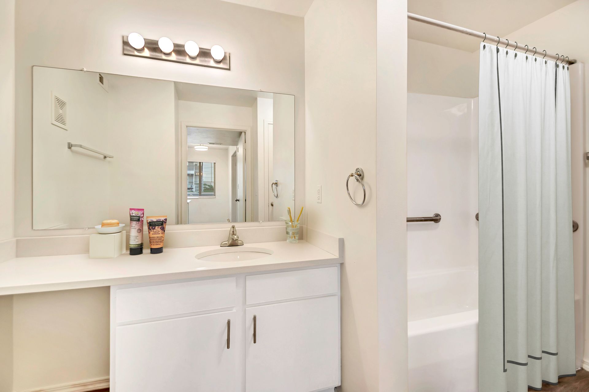 Bright bathroom vanity with mirror lights, white cabinets, and shower curtain beside bathtub at Creekside Village apartments in Sandy, UT.