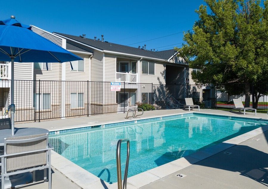 Apartment pool with clear water, blue umbrella, and lounge chairs beside beige buildings under a sunny sky at Creekside Village apartments in Sandy, UT.
