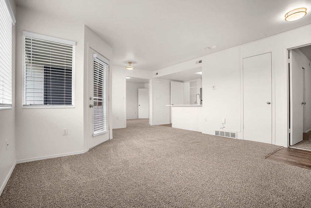 Bright empty living room with carpet, white walls, windows, and a kitchen pass-through at Creekside Village apartments in Sandy, UT.