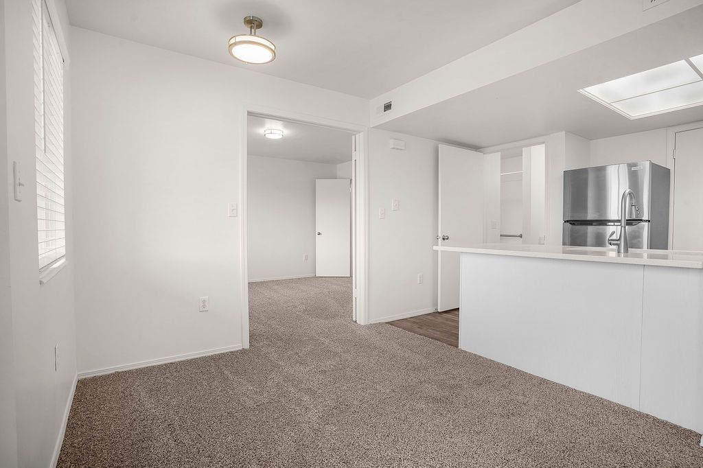Empty carpeted living room with white walls, open kitchen, and stainless steel appliances at Creekside Village apartments in Sandy, UT.