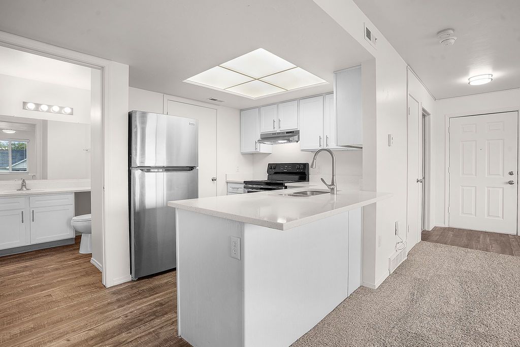 Bright modern kitchen with white cabinets, stainless steel fridge, and adjacent carpeted hallway and bathroom at Creekside Village apartments in Sandy, UT.