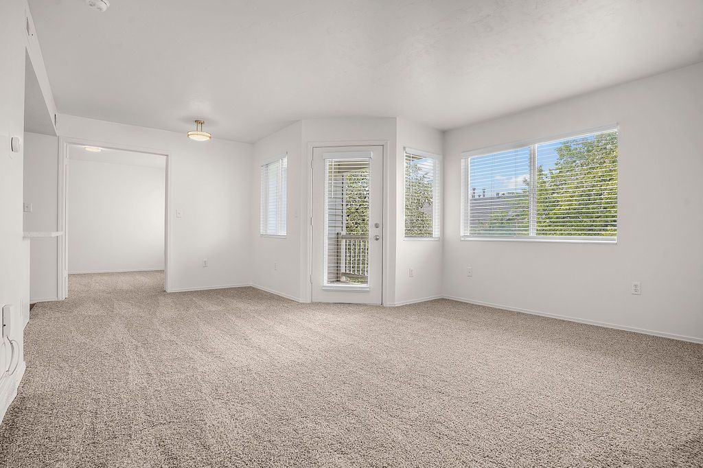 Empty carpeted living room with white walls, large windows, and a glass door opening to a balcony at Creekside Village apartments in Sandy, UT.