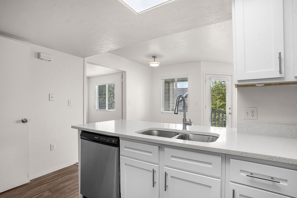 Bright white kitchen with island, stainless dishwasher, cabinets, and windows overlooking greenery at Creekside Village apartments in Sandy, UT.