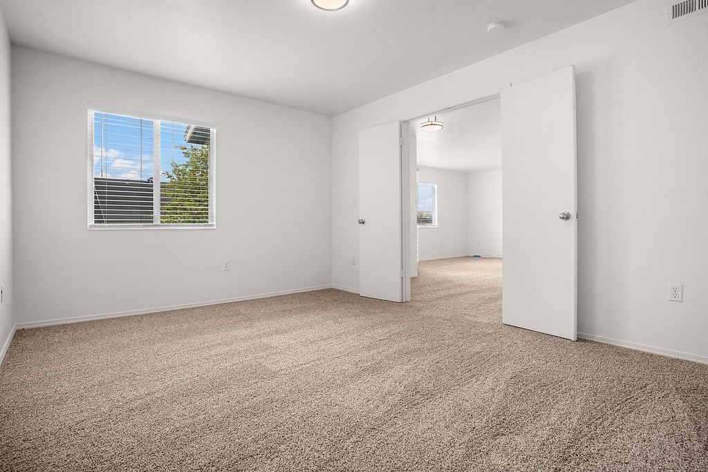 Empty carpeted bedroom with white walls, window, and open doorway to a bathroom or closet at Creekside Village apartments in Sandy, UT.