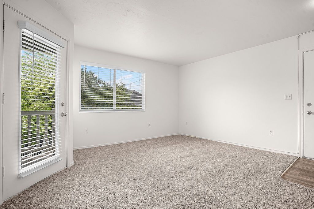 Empty carpeted room with white walls, window, and glass door to balcony at Creekside Village apartments in Sandy, UT.