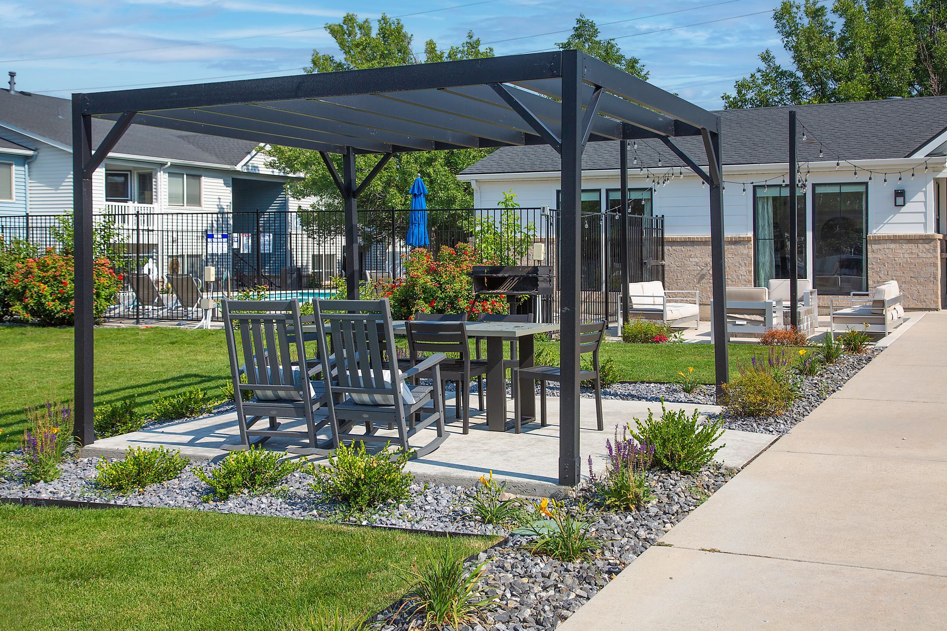 Outdoor patio with a black pergola, tables and chairs, and flower beds beside a building.
