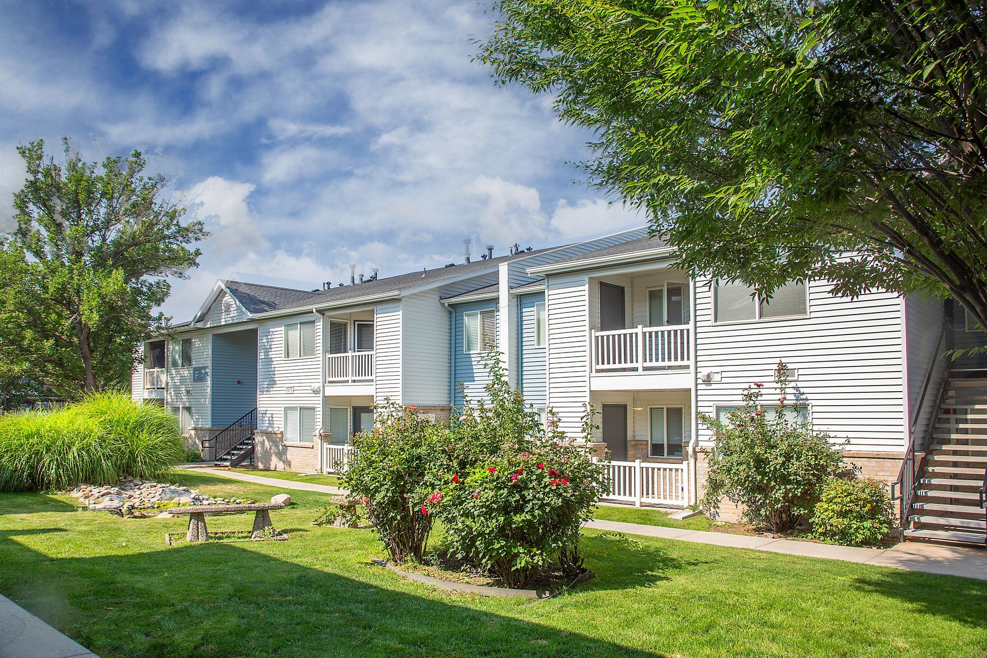 Apartment complex with white siding, green lawn, shrubs, and trees under a partly cloudy sky.