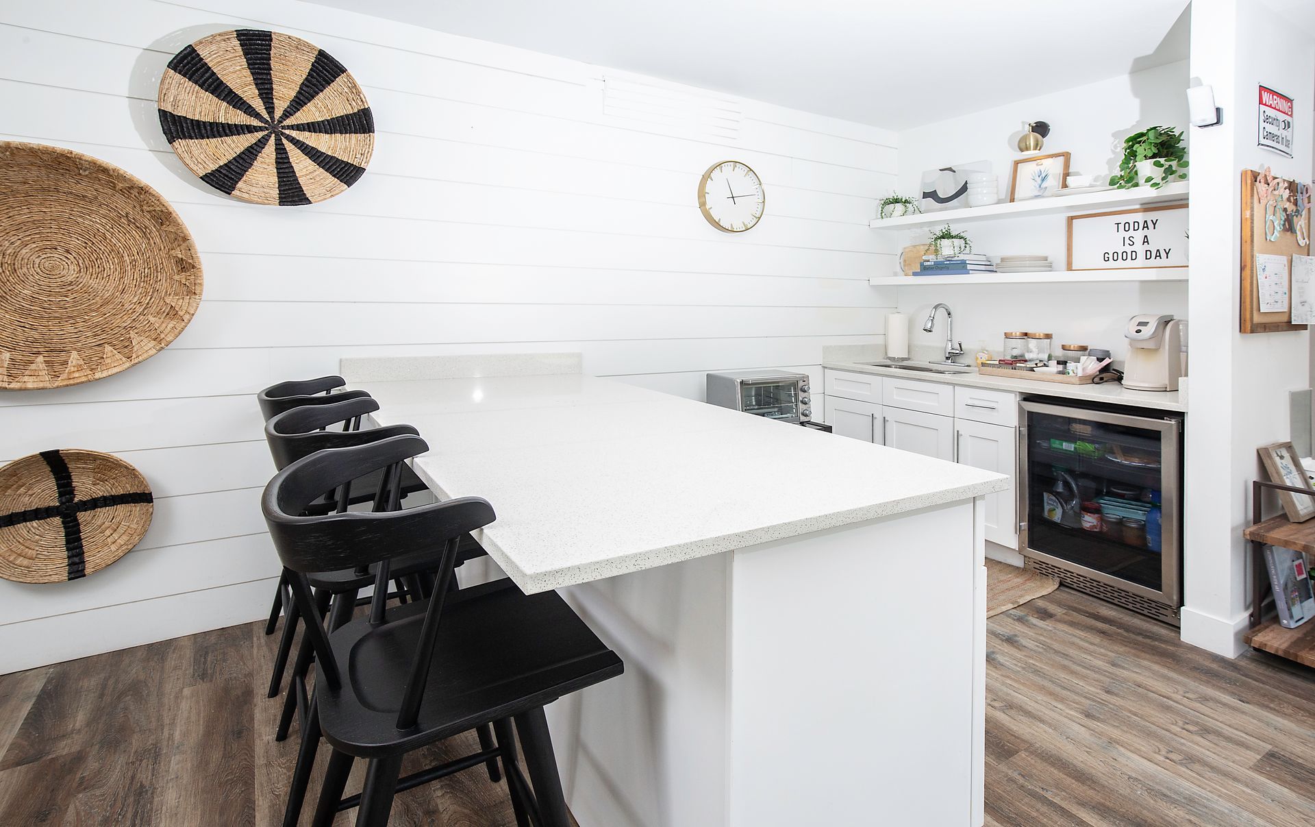 Bright white kitchen with island, black stools, woven wall decor, and shelves by a small fireplace.