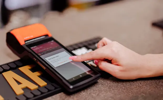 Hand holding a smartphone on a countertop beside an orange payment terminal and a black card reader