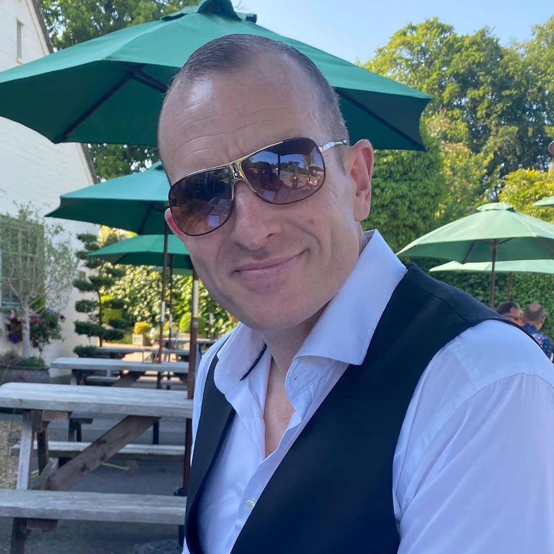 Smiling person in sunglasses at an outdoor picnic table under green umbrellas on a sunny day