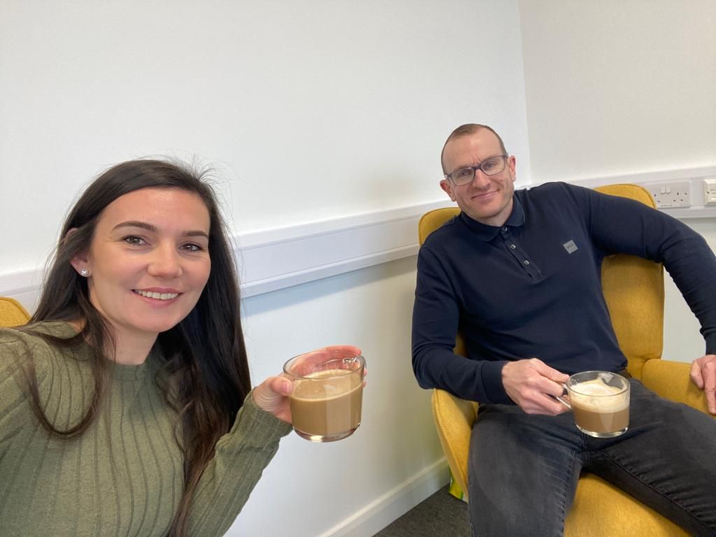 Two people seated indoors smiling and holding coffee in a bright room