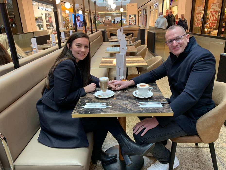 Two people seated at a cafe table in a shopping mall, smiling with coffee cups and desserts.
