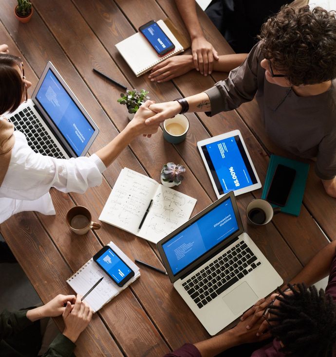 People around a wooden table shaking hands, with laptops, tablets, notebooks, and coffee cups.