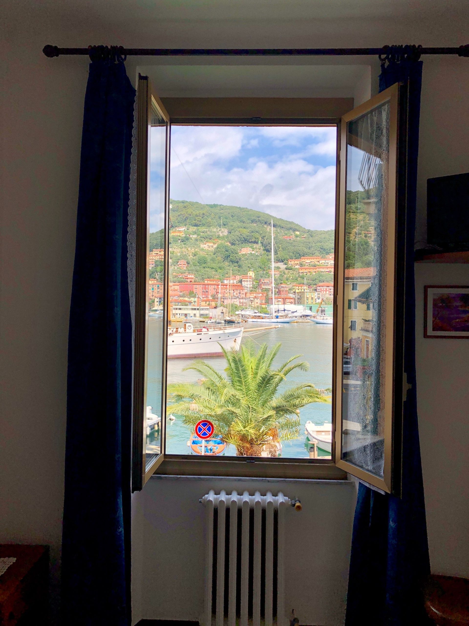 Finestra aperta con tende scure che si affaccia su un porto soleggiato e su una verde collina.