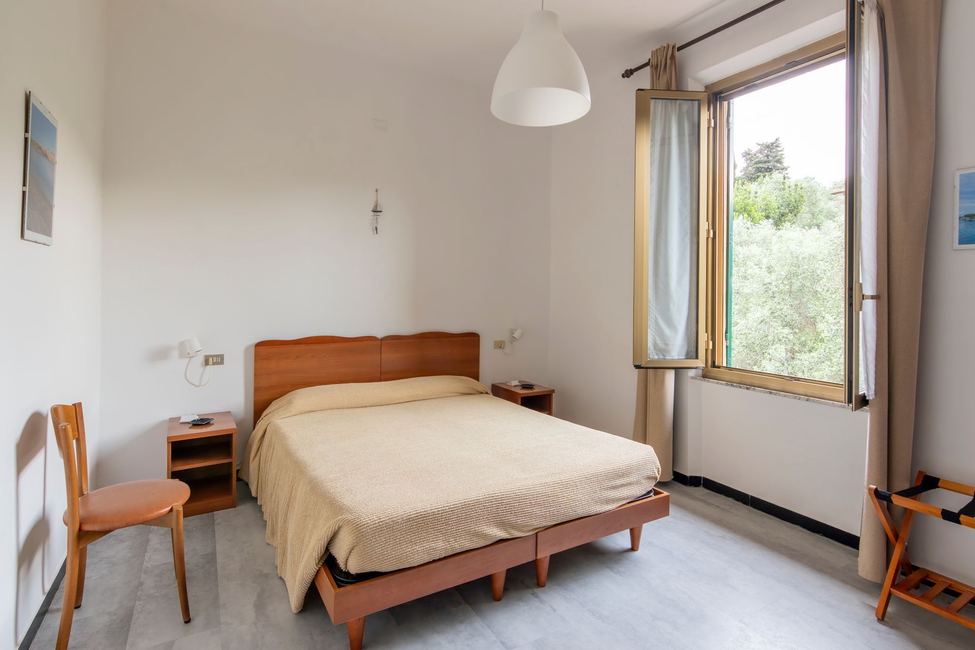 Camera da letto luminosa con letto in legno, biancheria da letto beige, sedia e finestra aperta con vista sul verde.