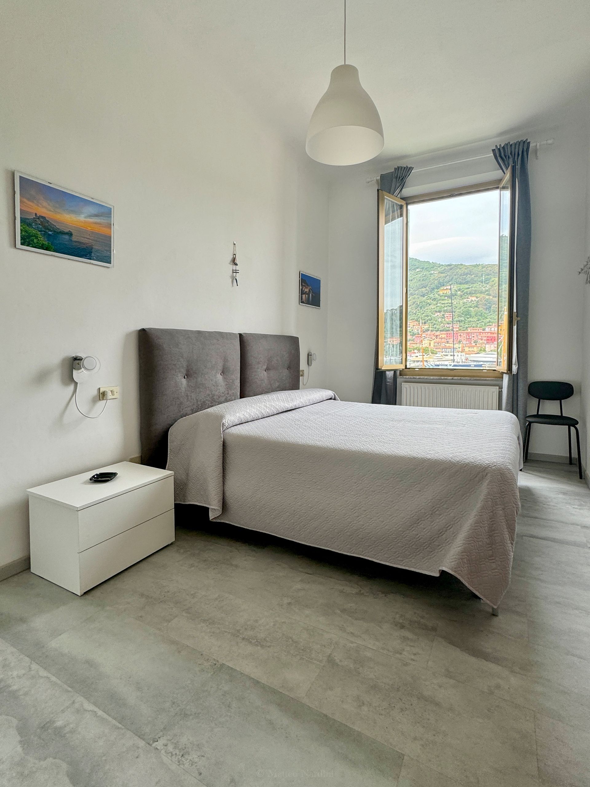 Camera da letto luminosa con letto grigio, pareti bianche, ampia finestra e vista sulla città.