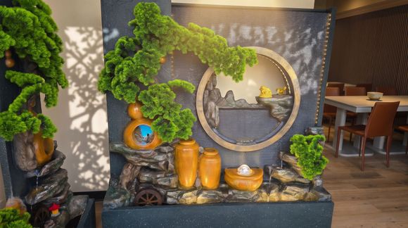 Decorative indoor fountain with bonsai, golden circular sculpture, and orange glass vases on a black stone table