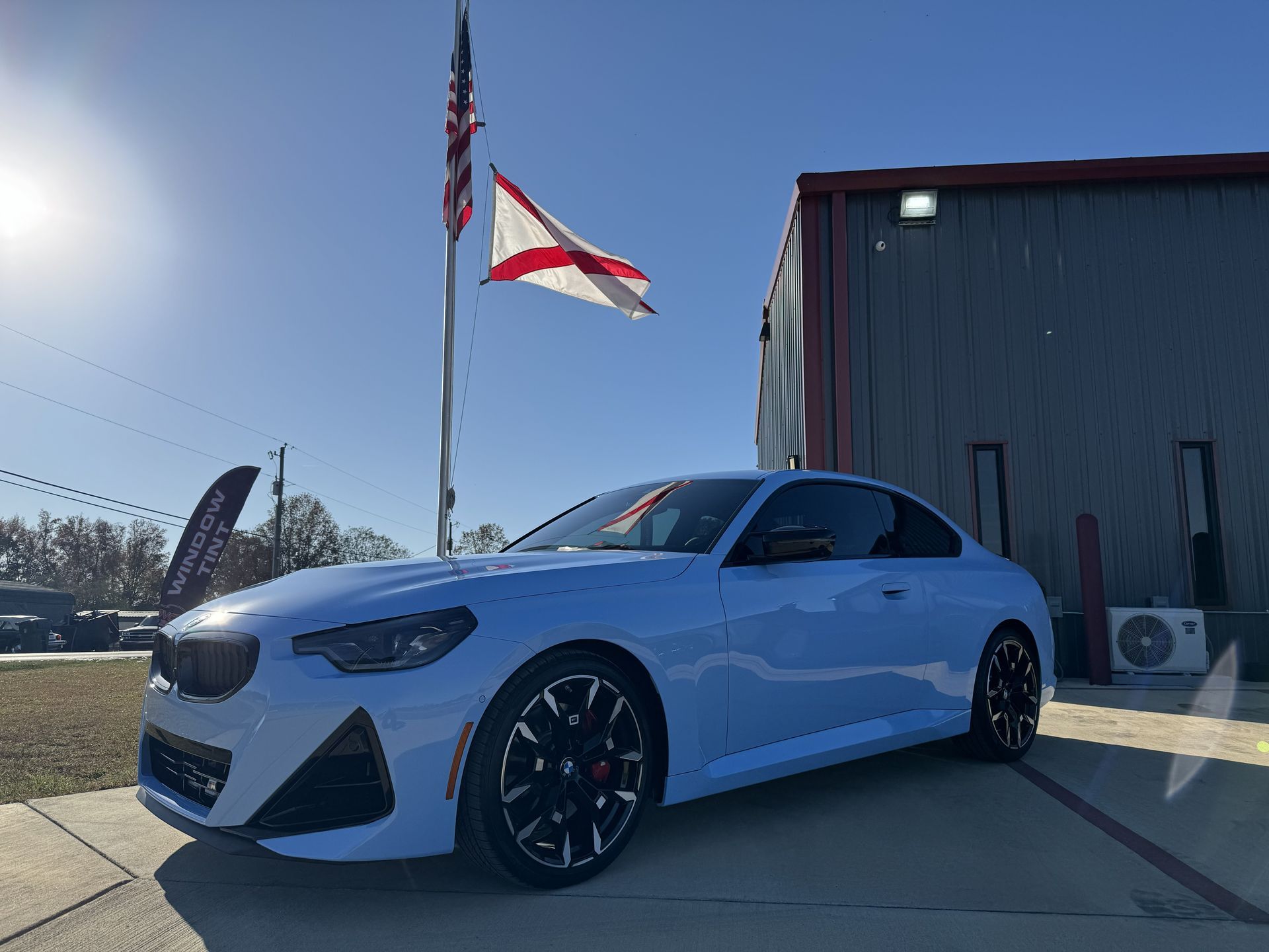 White sports coupe parked outside a building with two flags flying overhead in bright sunlight