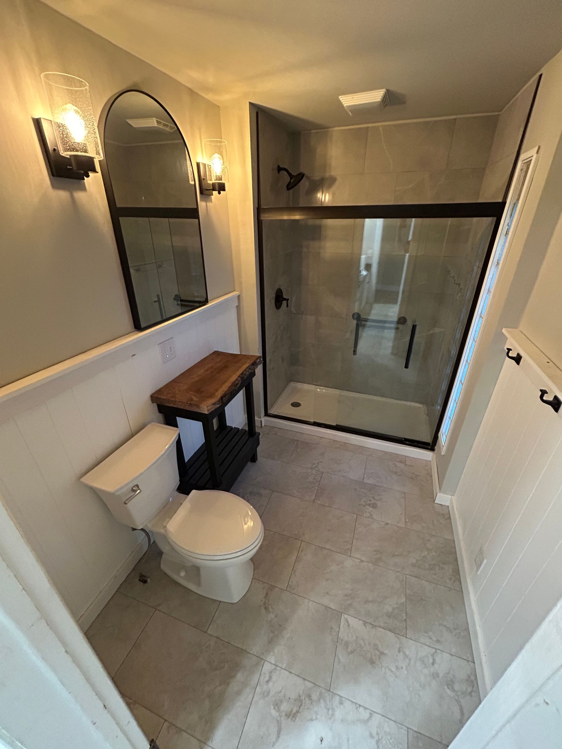 Small bathroom with toilet, wooden shelf, wall mirror, and glass shower enclosure.