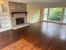 Empty living room with dark hardwood floors, brick fireplace, and large windows overlooking greenery