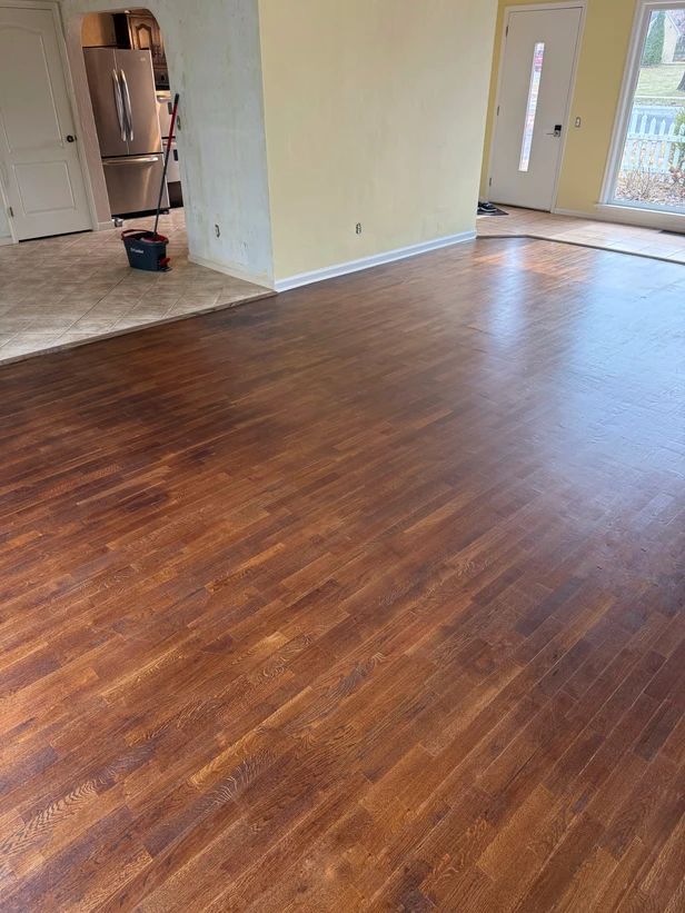 Empty room with dark wood floor, light walls, and a kitchen visible through an open doorway.