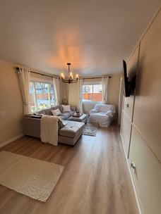 Bright living room with beige sofa, armchairs, large windows, and wall-mounted TV on the right.