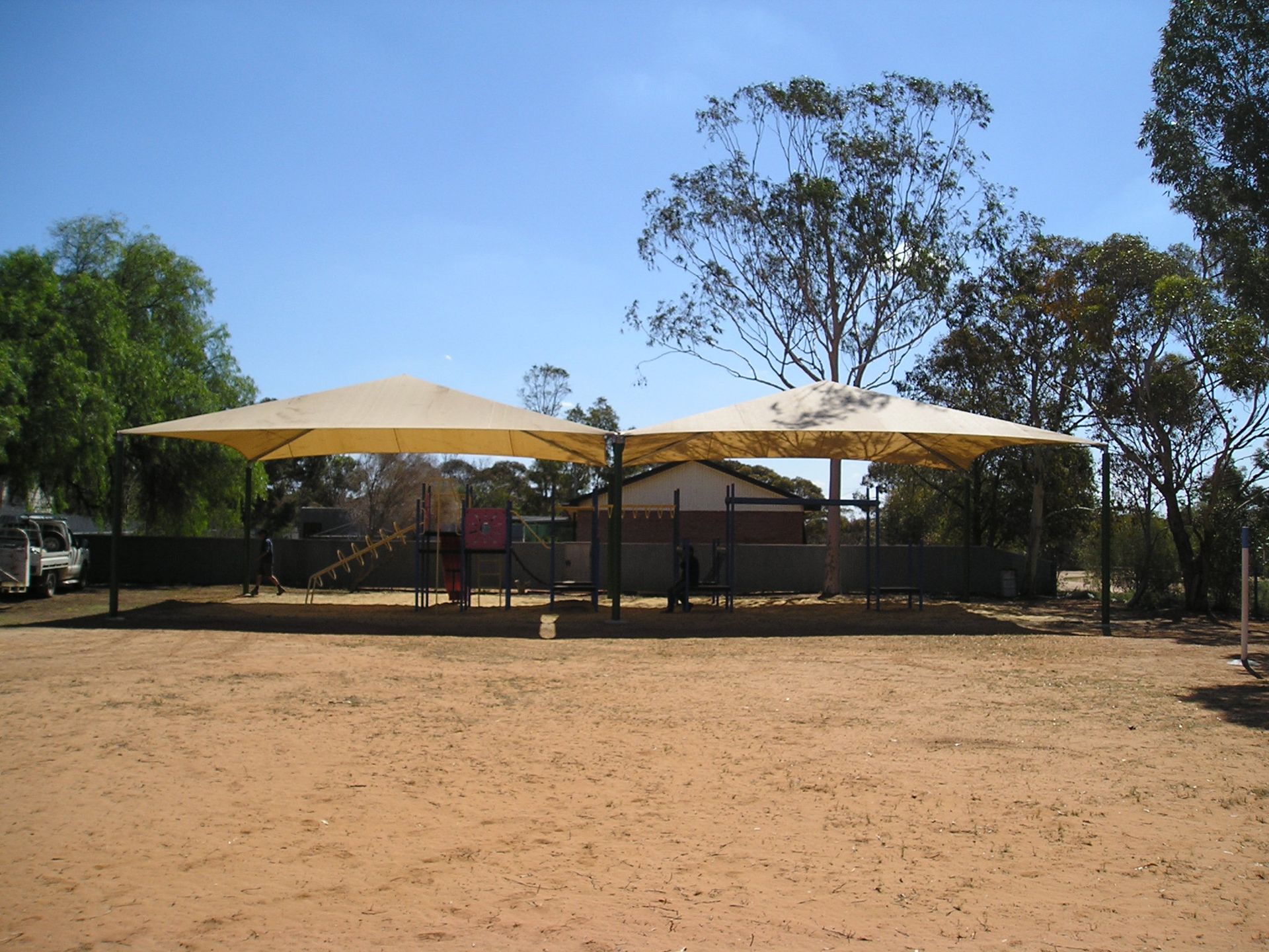 Outdoor park shelter with beige shade sails over a fenced area, trees and open dirt ground under a clear blue sky  — The Gazebo and Shade Centre in Jindera, NSW