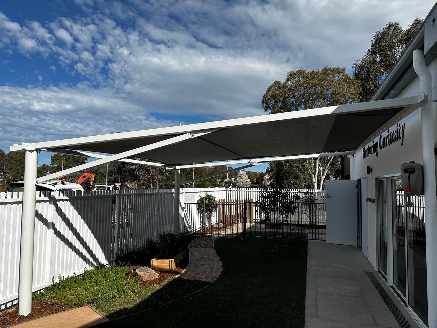 Covered outdoor patio beside a white building, with a fenced walkway and partly cloudy sky — The Gazebo and Shade Centre in Wodonga, VIC