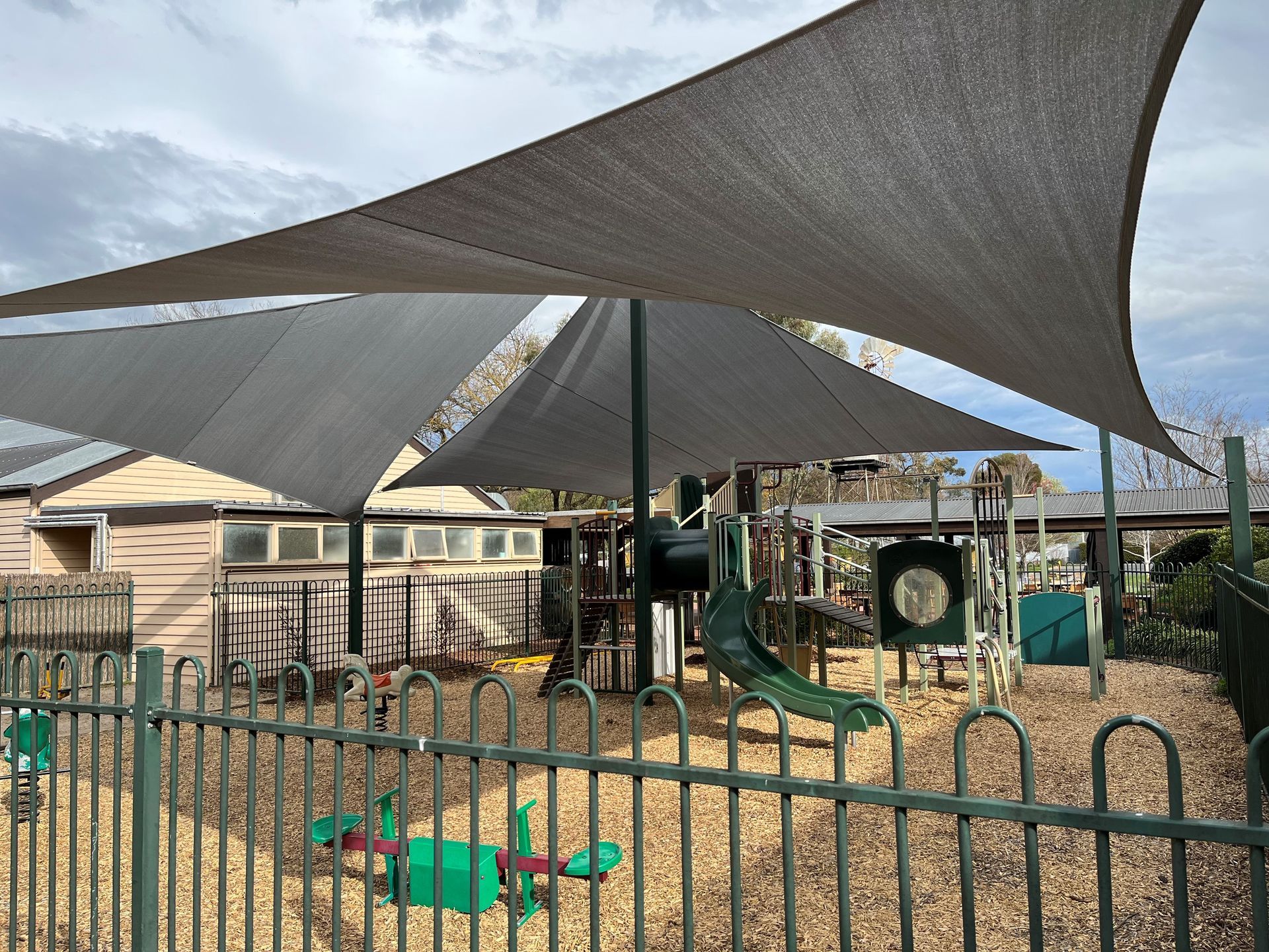 Shaded playground with green climbing equipment and a black fence in front of buildings   — The Gazebo and Shade Centre in Jindera, NSW