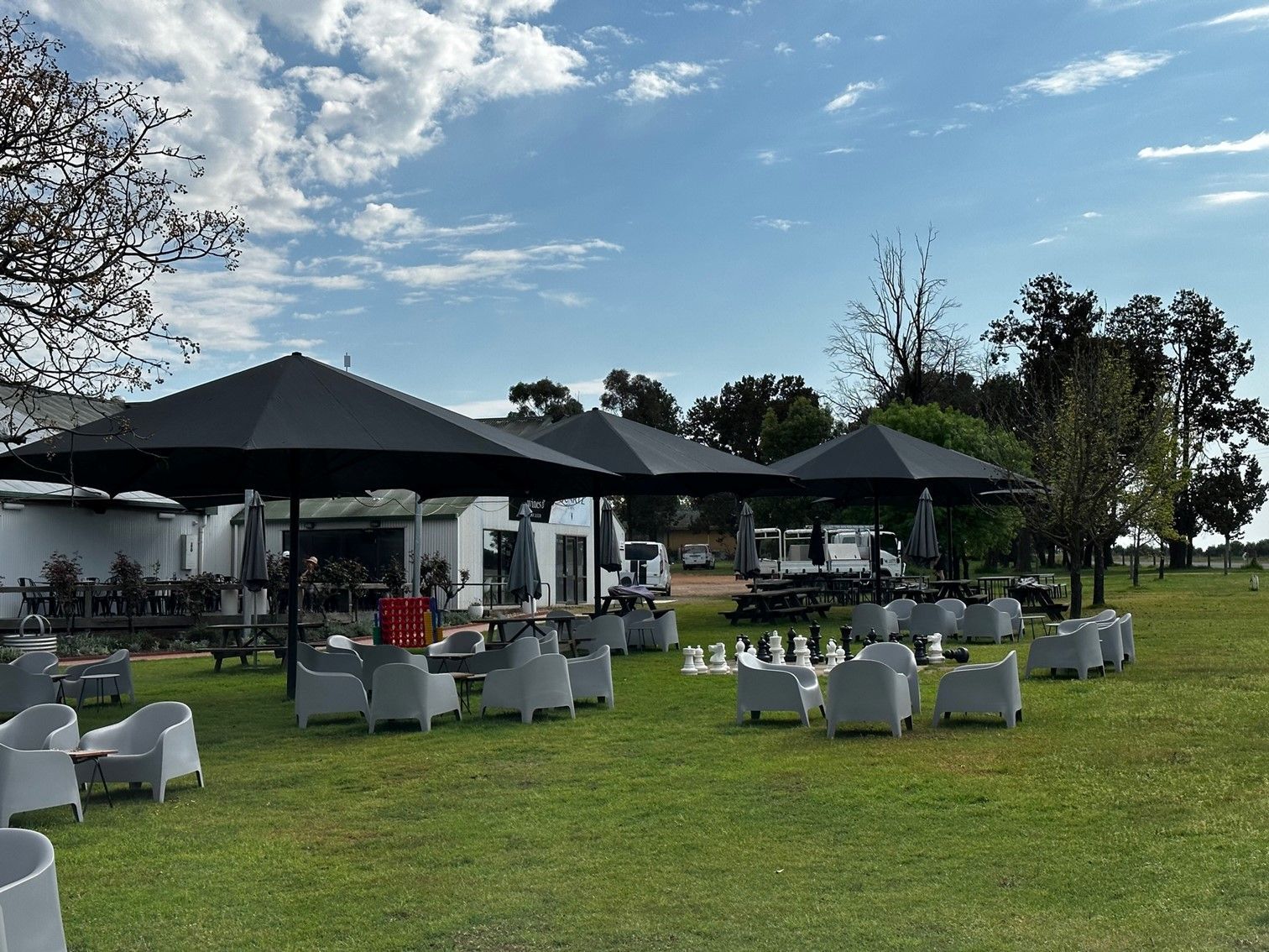 Outdoor event seating with white chairs around tables on a grassy lawn under black umbrellas near a lake — The Gazebo and Shade Centre in Yarrawonga, VIC
