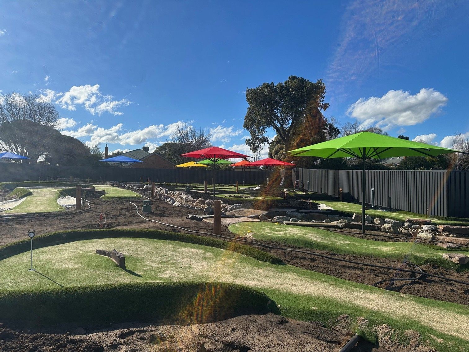Sunny mini golf course with colourful umbrellas and a fenced seating area under a blue sky  — The Gazebo and Shade Centre in Yarrawonga, VIC