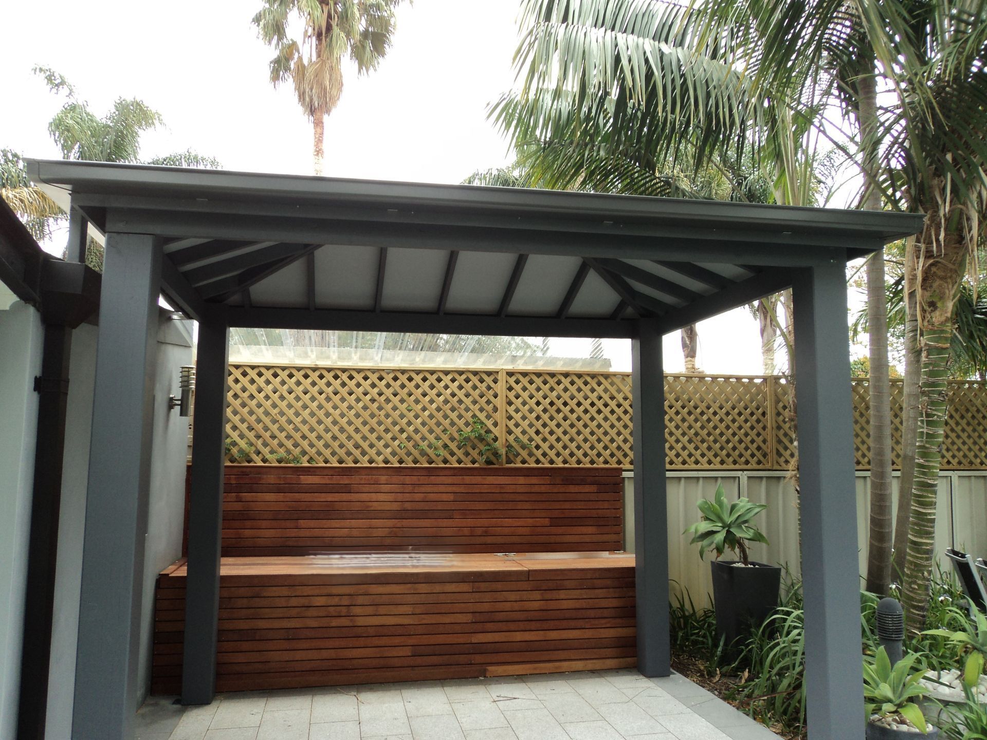 Covered outdoor patio with wood bar counter, lattice fence, and tropical plants under palm trees — The Gazebo and Shade Centre in Jindera, NSW
