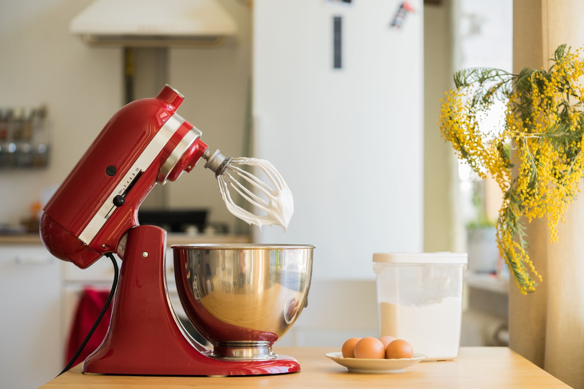 Red stand mixer whisking batter in a kitchen beside eggs, a measuring cup, and flowers.