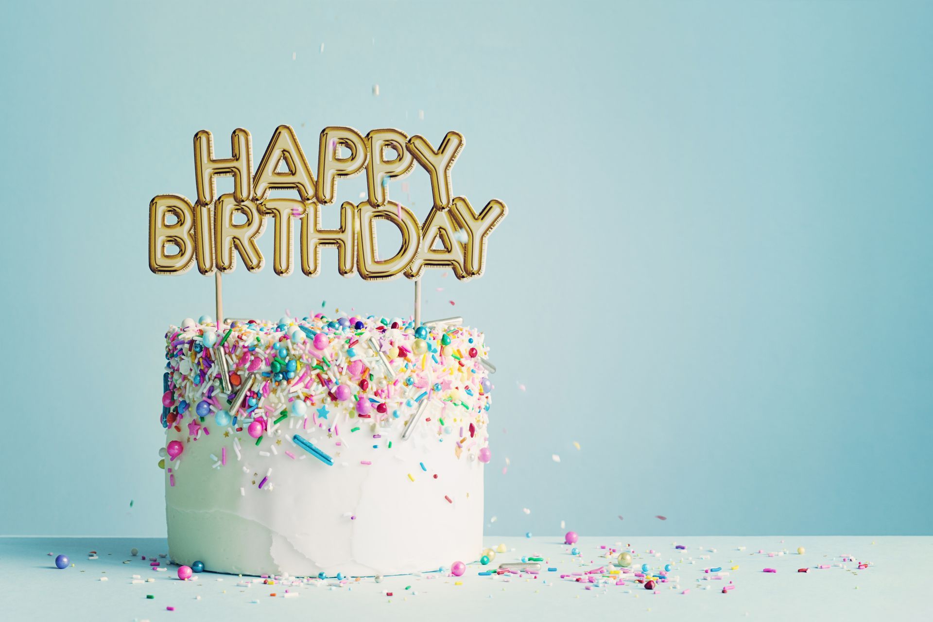Birthday cake with colorful sprinkles and gold “Happy Birthday” topper on a pastel blue background