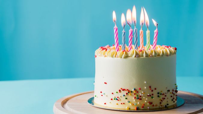 White frosted birthday cake with lit candles and colorful sprinkles on a plate against a blue background White frosted birthday cake with lit candles and colorful sprinkles on a plate against a blue background