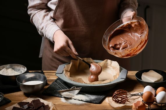 Baker pouring chocolate batter into a cake pan on a flour-dusted kitchen table with eggs and bowls. Baker pouring chocolate batter into a cake pan on a flour-dusted kitchen table with eggs and bowls.