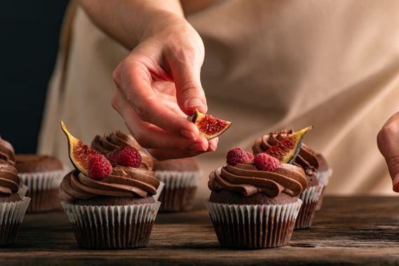 Hand placing fig slice on chocolate cupcakes topped with raspberries on a rustic table Hand placing fig slice on chocolate cupcakes topped with raspberries on a rustic table