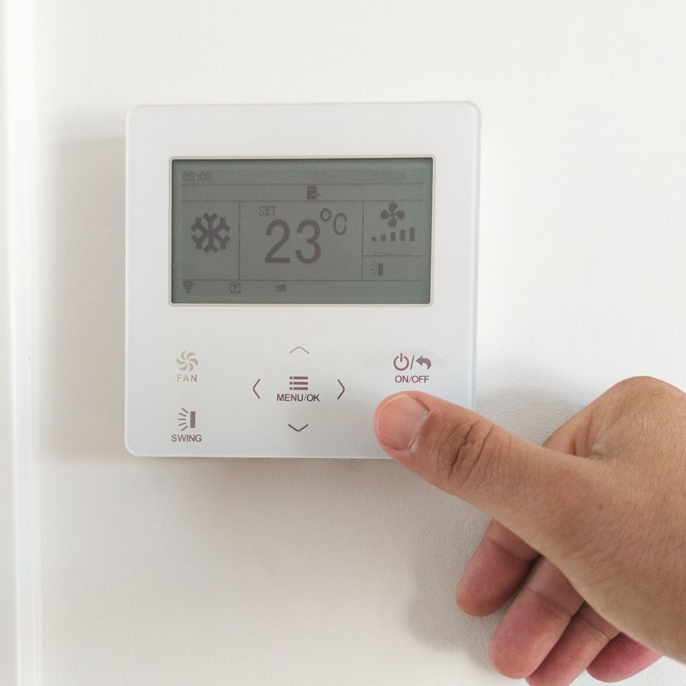 Hand pressing on/off button of a white thermostat displaying 23°C and snowflake icon, mounted on a white wall. Hand pressing on/off button of a white thermostat displaying 23°C and snowflake icon, mounted on a white wall.