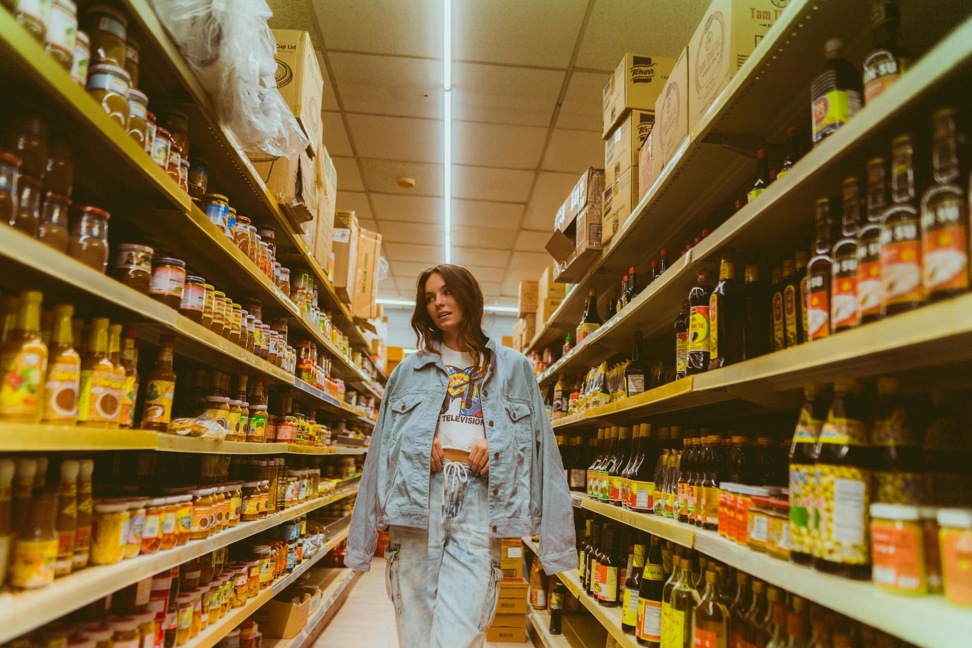 Person in a grocery store aisle lined with snacks and jars, standing beneath warm overhead lights.