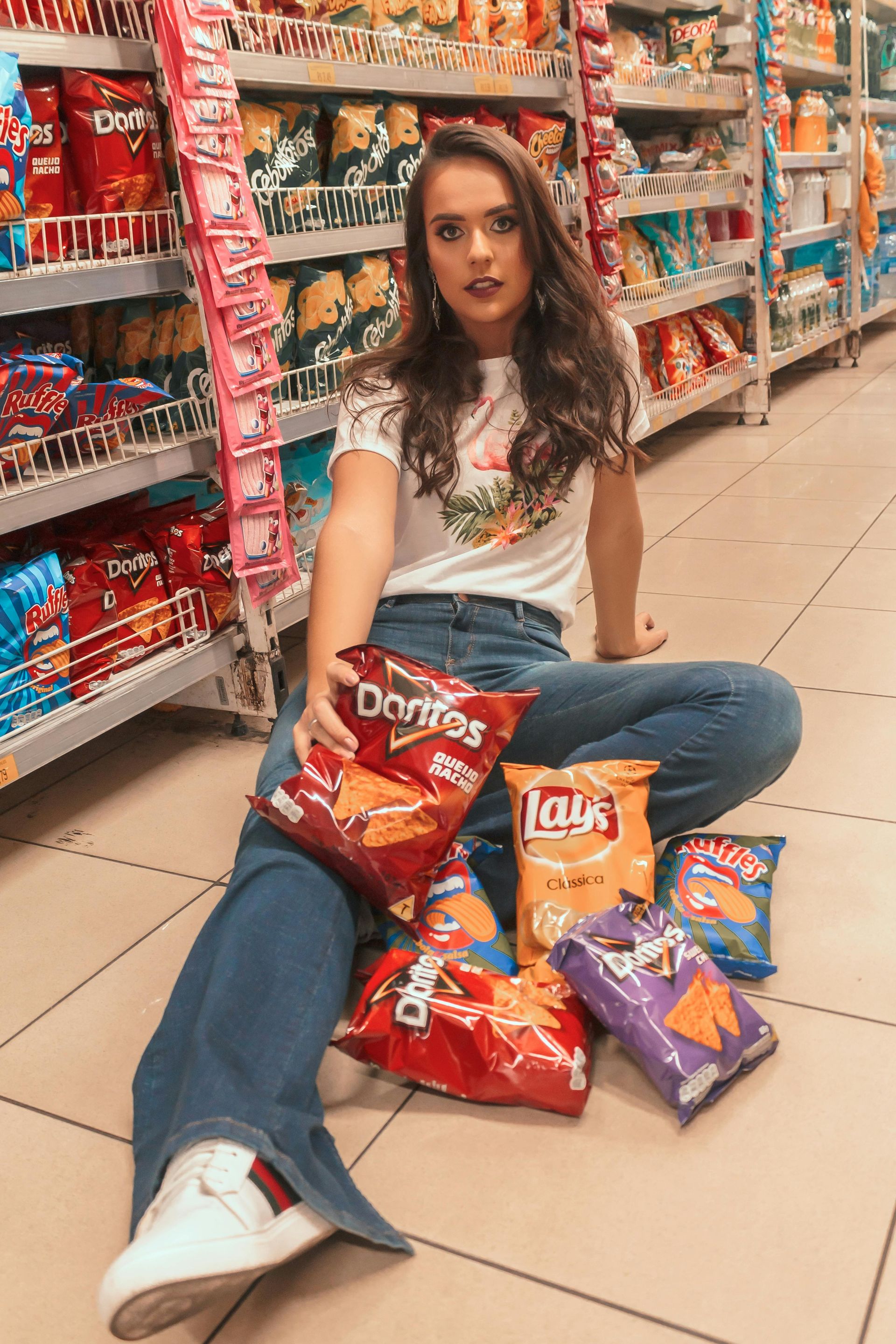 Woman sitting on grocery store floor surrounded by colorful snack bags, looking at the camera.