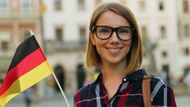 Smiling person with glasses holding a German flag outdoors in front of a blurred building