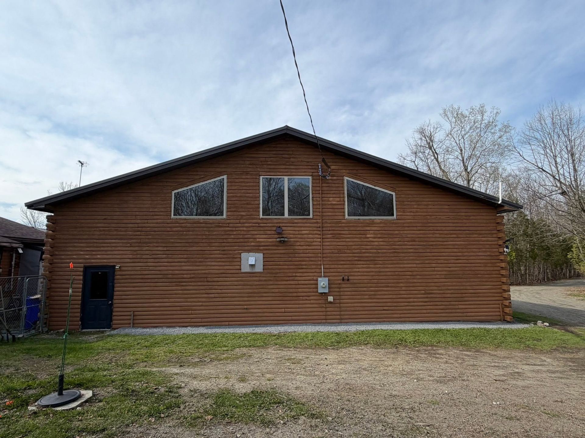 Wood-sided building with three windows, a black door, and a gravel yard under a cloudy sky