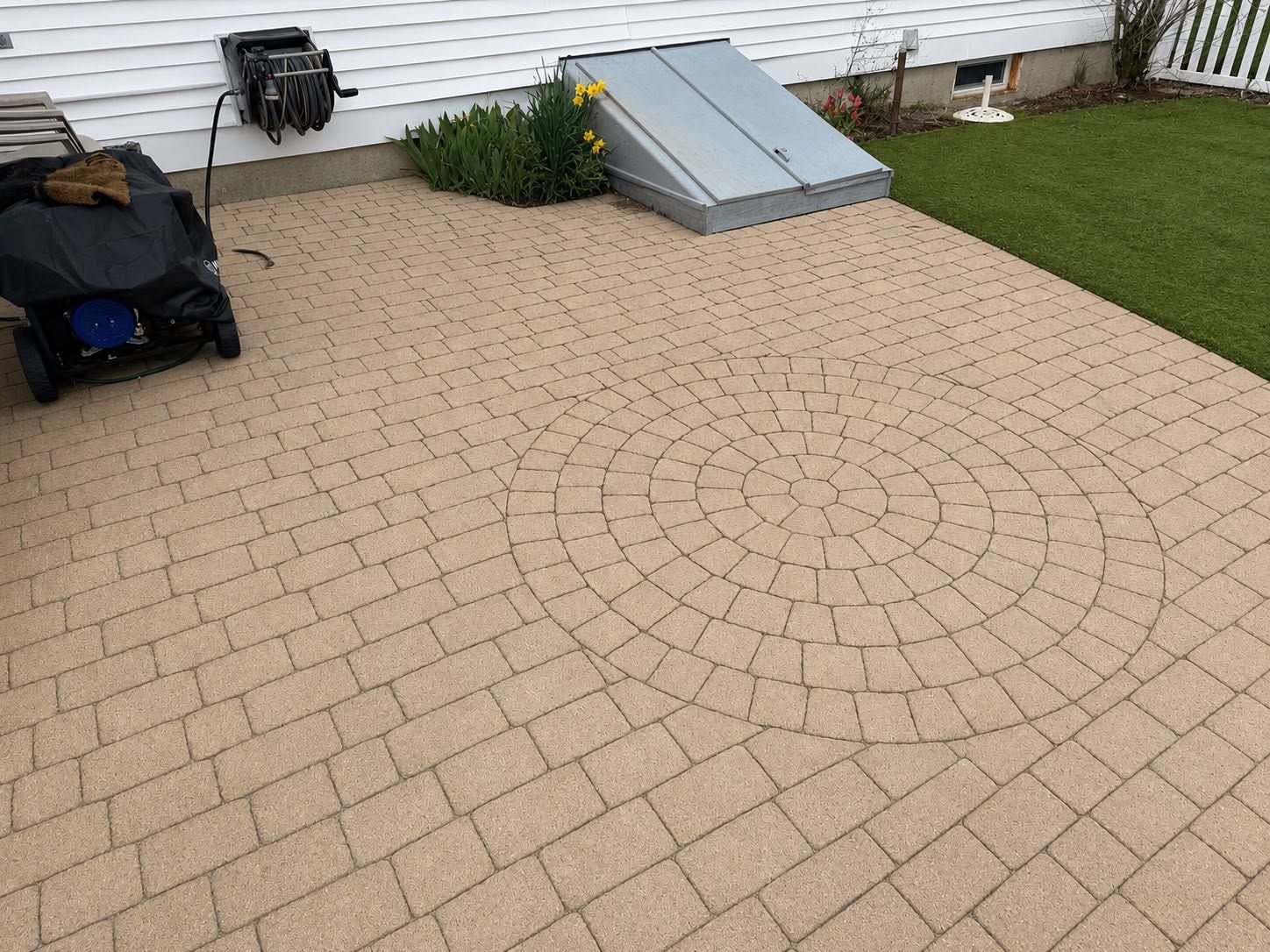 Paver patio beside a house with a gray basement hatch and outdoor tools